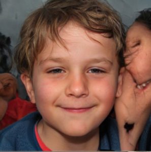 A small boy looks directly at the camera with a self-satisfied smile. Something very strange that looks like it is made of flesh, teeth and hair is on his right.