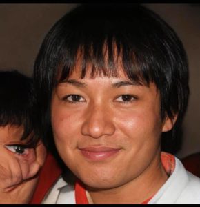 A smiling man looks direct at camera. What appears to be a fist with an eye looks over his right shoulder.