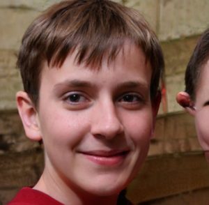 A young boy looks direct to camera, smiling slightly. To his right is a misshapen head with one ear sticking out like a cup handle.