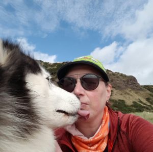 A white-faced husky in exterme close-up, licking the author's chin. The author is visible from the shoulders up, and is wearing dark glasses. The photo was taken outside with vegetated cliffs in the background under a partly cloudy blue sky.